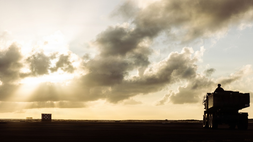 A dramatic silhouette photograph taken at what appears to be a military airfield or airstrip during golden hour or sunset. A large military vehicle with a raised launcher or equipment system is visible in the right foreground, while a checkered runway distance marker is visible in the middle distance. The scene is dominated by a moody, cloud-filled sky with rays of light breaking through.