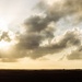 A dramatic silhouette photograph taken at what appears to be a military airfield or airstrip during golden hour or sunset. A large military vehicle with a raised launcher or equipment system is visible in the right foreground, while a checkered runway distance marker is visible in the middle distance. The scene is dominated by a moody, cloud-filled sky with rays of light breaking through.