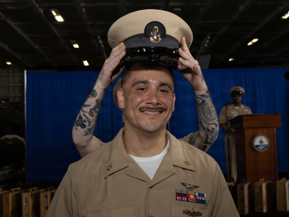 A U.S. Navy sailor is smiling as a chief petty officer cover (hat) is being placed on his head, indicating a chief petty officer pinning or advancement ceremony. He is wearing khaki service uniform with ribbons, warfare devices, and collar insignia. The ceremony appears to be taking place in a ship's hangar bay with a blue curtain backdrop and a podium visible in the background.