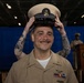 A U.S. Navy sailor is smiling as a chief petty officer cover (hat) is being placed on his head, indicating a chief petty officer pinning or advancement ceremony. He is wearing khaki service uniform with ribbons, warfare devices, and collar insignia. The ceremony appears to be taking place in a ship's hangar bay with a blue curtain backdrop and a podium visible in the background.