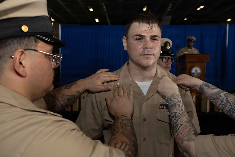A military promotion or pinning ceremony is taking place, with two individuals affixing insignia or rank devices to the collar/shoulders of a young service member in a tan/khaki uniform. The setting appears to be an official military venue with a blue curtain backdrop and a podium visible in the background. Additional uniformed personnel are present in the background observing the ceremony.