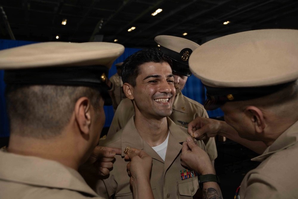 A military promotion or pinning ceremony is taking place, with two uniformed personnel pinning insignia onto the collar of a smiling service member in the center. The central figure appears to be receiving a new rank or qualification device, indicated by the hands of colleagues attaching items to both sides of his collar. The setting appears to be aboard a naval vessel, likely in a hangar bay or similar large interior space.