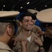 A military promotion or pinning ceremony is taking place, with two uniformed personnel pinning insignia onto the collar of a smiling service member in the center. The central figure appears to be receiving a new rank or qualification device, indicated by the hands of colleagues attaching items to both sides of his collar. The setting appears to be aboard a naval vessel, likely in a hangar bay or similar large interior space.