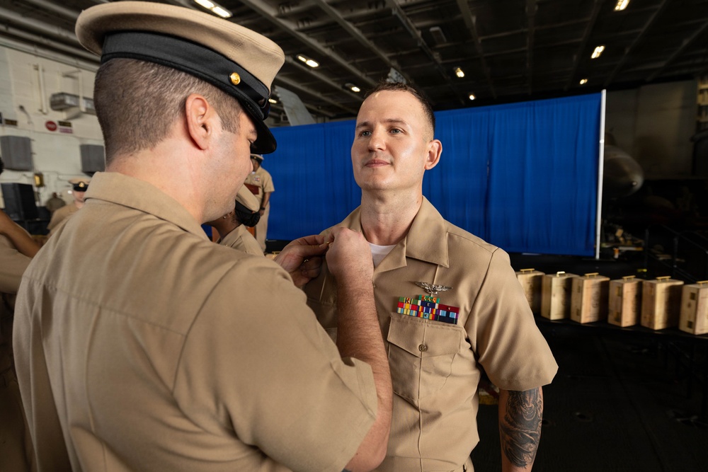 A military ceremony is taking place in what appears to be a ship's hangar bay, where one uniformed service member is pinning a medal or insignia onto another. The recipient is wearing a tan/khaki Navy uniform with existing ribbons, a warfare device, and a visible tattoo on his left arm. The setting and uniforms are consistent with U.S. Navy personnel.