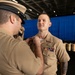 A military ceremony is taking place in what appears to be a ship's hangar bay, where one uniformed service member is pinning a medal or insignia onto another. The recipient is wearing a tan/khaki Navy uniform with existing ribbons, a warfare device, and a visible tattoo on his left arm. The setting and uniforms are consistent with U.S. Navy personnel.