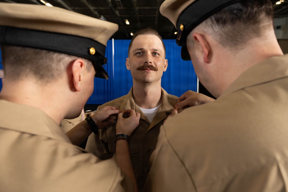 A military promotion or pinning ceremony is taking place, with two uniformed personnel pinning insignia onto the collar of a third service member. The central figure, wearing a tan/khaki Navy working uniform, stands at attention with a slight smile while two colleagues on either side attach rank devices or devices to his collar. The setting appears to be an indoor military venue with a blue curtain backdrop.