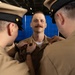 A military promotion or pinning ceremony is taking place, with two uniformed personnel pinning insignia onto the collar of a third service member. The central figure, wearing a tan/khaki Navy working uniform, stands at attention with a slight smile while two colleagues on either side attach rank devices or devices to his collar. The setting appears to be an indoor military venue with a blue curtain backdrop.