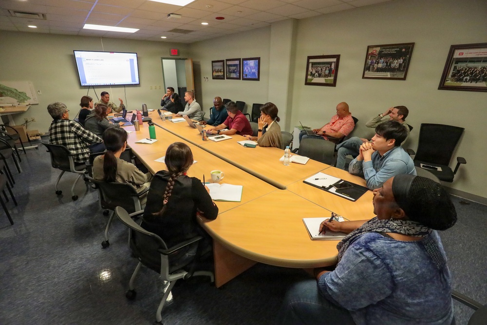 A professional meeting or briefing is taking place in a conference room with approximately 15 participants seated around a large oval wooden table. A presenter appears to be speaking at the front of the room near a wall-mounted display screen showing a presentation. The room is decorated with several framed photographs on the walls, suggesting an organizational or institutional setting.
