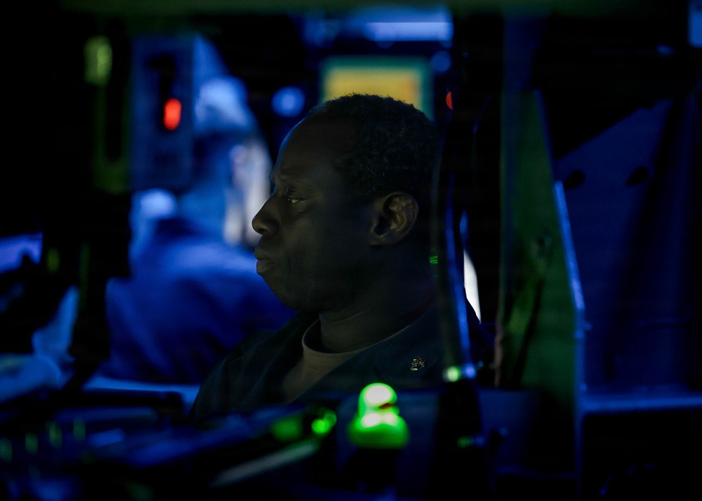 A person in what appears to be a military uniform is seated at a dimly lit operations console, likely aboard a naval vessel. The environment is characterized by low red and green indicator lights typical of a combat information center or ship's bridge. The dramatic low-key lighting creates a high-contrast profile view of the individual monitoring equipment.