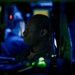 A person in what appears to be a military uniform is seated at a dimly lit operations console, likely aboard a naval vessel. The environment is characterized by low red and green indicator lights typical of a combat information center or ship's bridge. The dramatic low-key lighting creates a high-contrast profile view of the individual monitoring equipment.