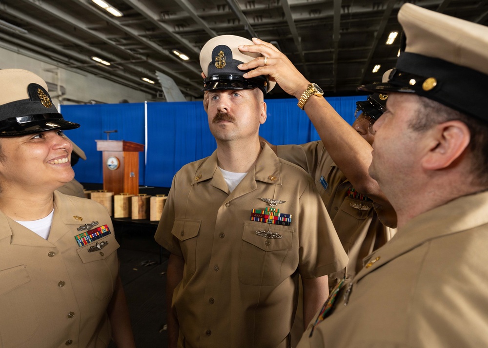 A U.S. Navy chief petty officer promotion or pinning ceremony is taking place in what appears to be a ship's hangar bay. A fellow sailor is placing a chief petty officer combination cover (white-topped cap with anchor insignia) on the head of a newly promoted chief, while another sailor looks on smiling. The setting includes a wooden podium with a Navy seal in the background, consistent with a formal military ceremony.