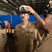 A U.S. Navy chief petty officer promotion or pinning ceremony is taking place in what appears to be a ship's hangar bay. A fellow sailor is placing a chief petty officer combination cover (white-topped cap with anchor insignia) on the head of a newly promoted chief, while another sailor looks on smiling. The setting includes a wooden podium with a Navy seal in the background, consistent with a formal military ceremony.