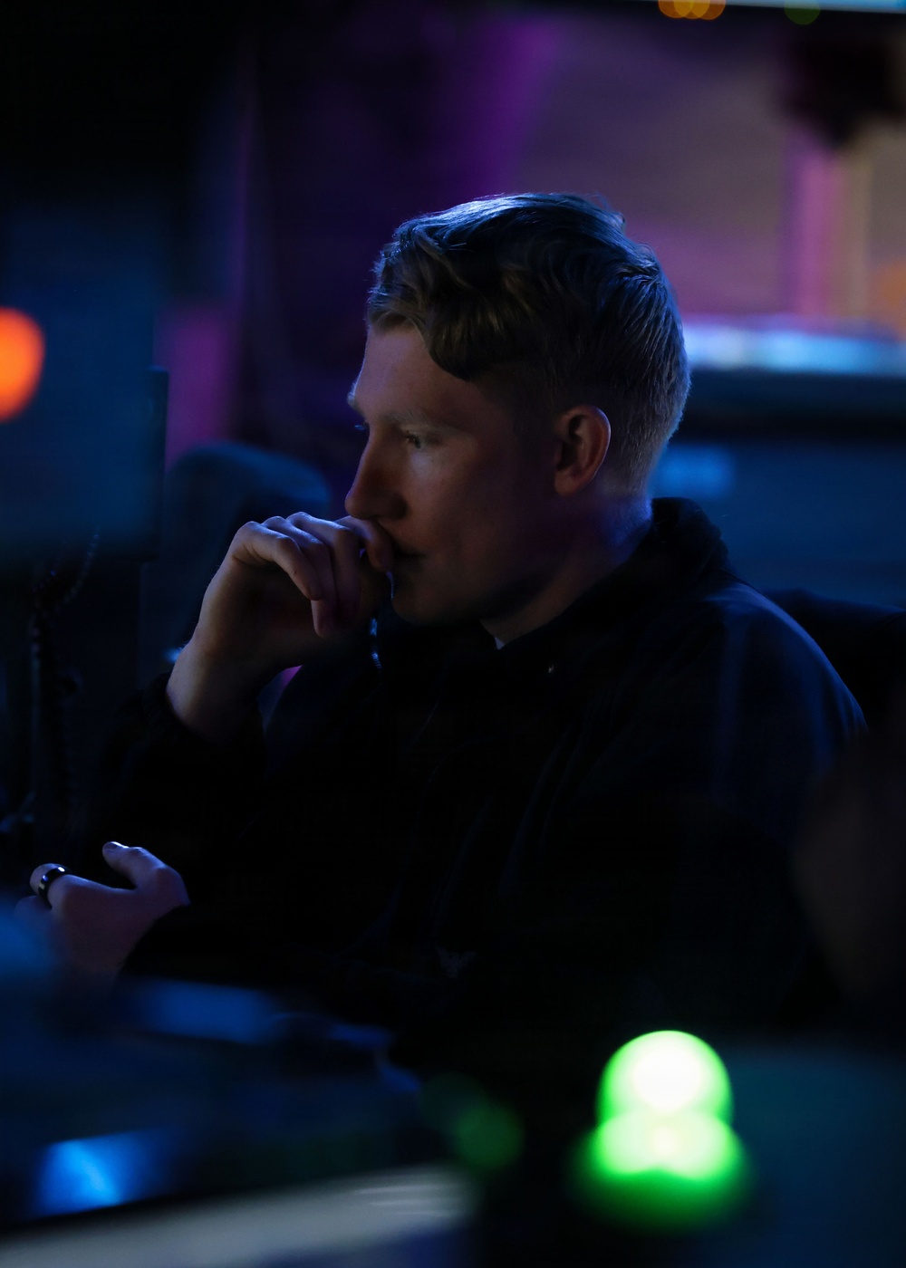 A person is seated in a dimly lit environment, likely a control room, studio, or entertainment venue, illuminated by blue and purple ambient lighting. They appear to be in a contemplative or focused pose with one hand raised to their chin. The scene is characterized by low-key dramatic lighting with visible indicator lights in the foreground.