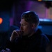 A person is seated in a dimly lit environment, likely a control room, studio, or entertainment venue, illuminated by blue and purple ambient lighting. They appear to be in a contemplative or focused pose with one hand raised to their chin. The scene is characterized by low-key dramatic lighting with visible indicator lights in the foreground.