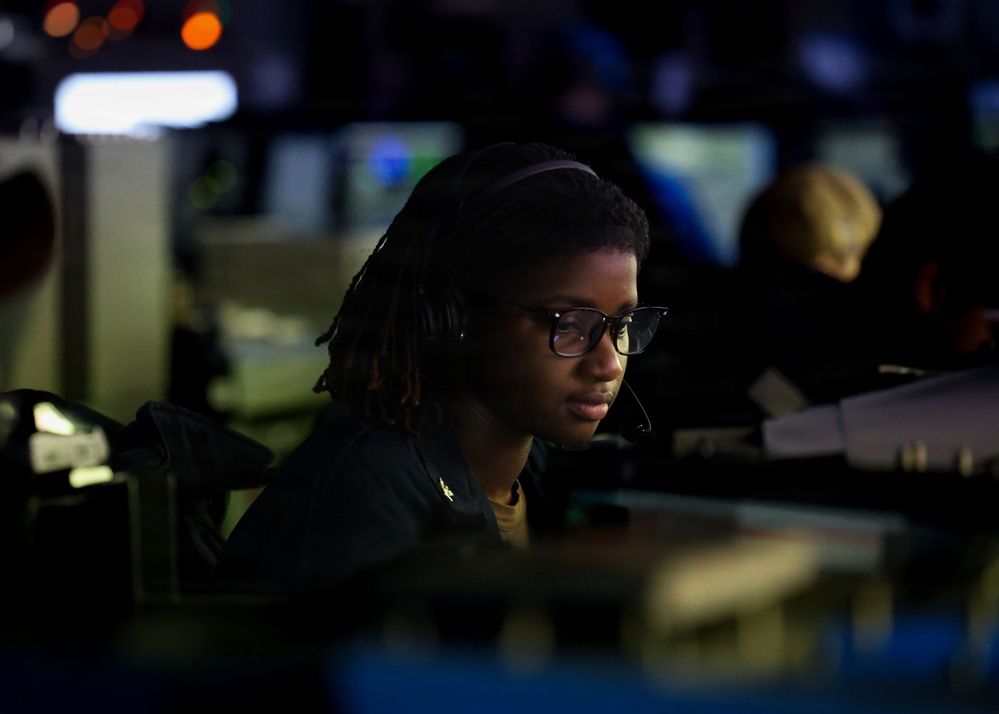 A person wearing glasses, a headset, and a dark uniform is seated at a workstation in a dimly lit operations or command center environment. The individual appears focused on screens or equipment in front of them. Multiple monitors and other personnel are visible in the blurred background.