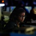 A person wearing glasses, a headset, and a dark uniform is seated at a workstation in a dimly lit operations or command center environment. The individual appears focused on screens or equipment in front of them. Multiple monitors and other personnel are visible in the blurred background.