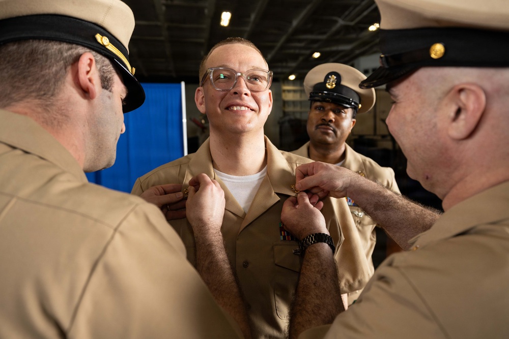 A U.S. Navy promotion or pinning ceremony is taking place, with two senior enlisted personnel simultaneously pinning insignia onto the collar of a smiling sailor in the center. The recipient is wearing a khaki uniform and appears pleased, suggesting this is a rank advancement ceremony. A third sailor in the background observes the proceedings.
