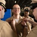 A U.S. Navy promotion or pinning ceremony is taking place, with two senior enlisted personnel simultaneously pinning insignia onto the collar of a smiling sailor in the center. The recipient is wearing a khaki uniform and appears pleased, suggesting this is a rank advancement ceremony. A third sailor in the background observes the proceedings.