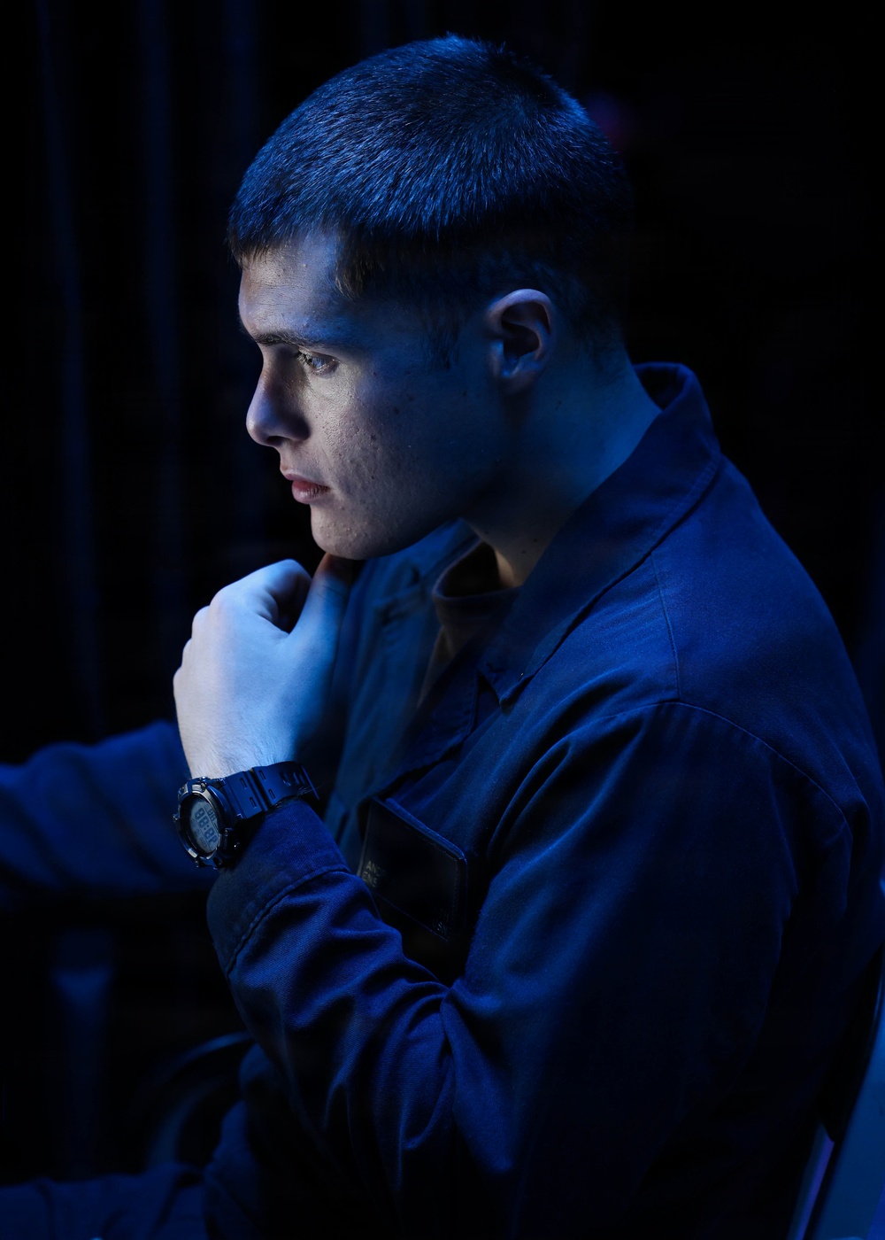 A young male subject is photographed in dramatic side-profile lighting with a strong blue color cast against a dark background. He is posed in a contemplative stance with his fist raised near his chin. The image appears to be a stylized portrait, possibly military or professional in context given his uniform-like attire.