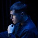A young male subject is photographed in dramatic side-profile lighting with a strong blue color cast against a dark background. He is posed in a contemplative stance with his fist raised near his chin. The image appears to be a stylized portrait, possibly military or professional in context given his uniform-like attire.