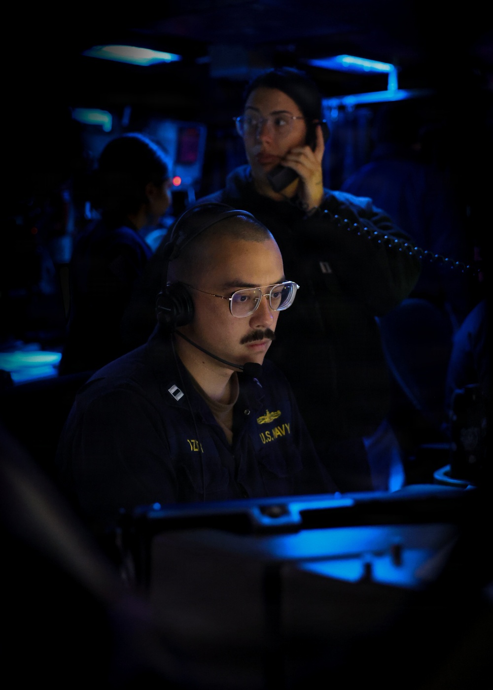 A U.S. Navy personnel member wearing a headset and clear-framed glasses is focused on a workstation console in a dimly lit combat information center or operations room aboard a naval vessel. Behind them, a second individual is speaking on a telephone handset. The environment is illuminated primarily by blue ambient lighting typical of shipboard tactical operations spaces.