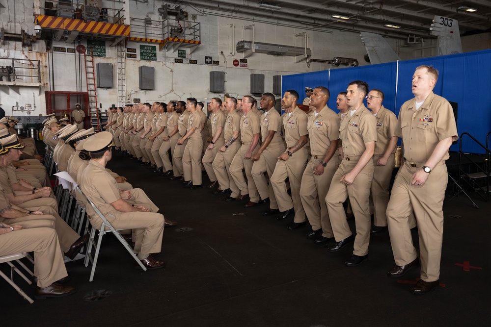 A formal military ceremony is taking place inside what appears to be a naval vessel's hangar bay, likely an aircraft carrier. A long row of U.S. Navy personnel in tan/khaki service uniforms stand at attention or in a recitation posture, facing a seated audience of fellow sailors. The setting, uniforms, and formation suggest this is a chief petty officer pinning ceremony or similar advancement/promotion event.