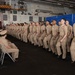 A formal military ceremony is taking place inside what appears to be a naval vessel's hangar bay, likely an aircraft carrier. A long row of U.S. Navy personnel in tan/khaki service uniforms stand at attention or in a recitation posture, facing a seated audience of fellow sailors. The setting, uniforms, and formation suggest this is a chief petty officer pinning ceremony or similar advancement/promotion event.