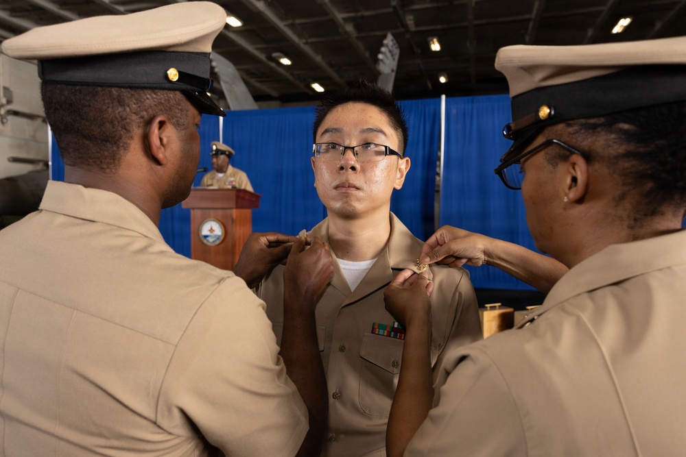 A military promotion or pinning ceremony is taking place, with two senior naval personnel in tan/khaki uniforms pinning insignia onto the collar of a junior sailor. The recipient, wearing glasses and a tan Navy uniform with service ribbons, stands at attention while the ceremony is conducted. A fourth individual in uniform is visible at a podium in the background, likely presiding over the ceremony.