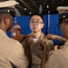 A military promotion or pinning ceremony is taking place, with two senior naval personnel in tan/khaki uniforms pinning insignia onto the collar of a junior sailor. The recipient, wearing glasses and a tan Navy uniform with service ribbons, stands at attention while the ceremony is conducted. A fourth individual in uniform is visible at a podium in the background, likely presiding over the ceremony.