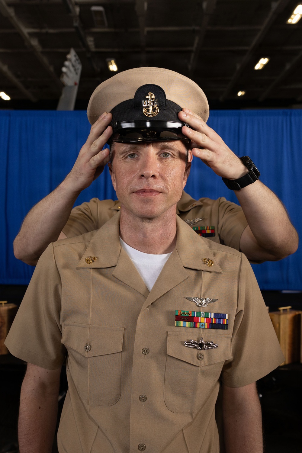 A U.S. Navy enlisted service member in khaki uniform is placing or adjusting a combination cover (cap) on their head. The individual displays multiple ribbons, warfare qualification badges, and collar devices consistent with a senior enlisted rank. The setting appears to be an indoor military venue with a blue curtain backdrop.