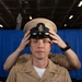 A U.S. Navy enlisted service member in khaki uniform is placing or adjusting a combination cover (cap) on their head. The individual displays multiple ribbons, warfare qualification badges, and collar devices consistent with a senior enlisted rank. The setting appears to be an indoor military venue with a blue curtain backdrop.