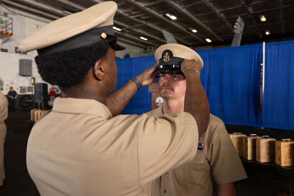 A U.S. Navy promotion ceremony is taking place in what appears to be a ship's hangar bay. One sailor in khaki uniform is placing a Chief Petty Officer combination cover (hat) onto another sailor's head, a traditional part of the CPO pinning/frocking ceremony. The recipient's cover displays a gold Chief Petty Officer anchor insignia, indicating advancement to the Chief Petty Officer rank.