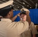 A U.S. Navy promotion ceremony is taking place in what appears to be a ship's hangar bay. One sailor in khaki uniform is placing a Chief Petty Officer combination cover (hat) onto another sailor's head, a traditional part of the CPO pinning/frocking ceremony. The recipient's cover displays a gold Chief Petty Officer anchor insignia, indicating advancement to the Chief Petty Officer rank.