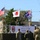 This image depicts a military ceremony for 'Orient Shield 2026,' a joint training exercise involving the United States, Japan, and Australia. Three soldiers are raising their respective national flags on a raised platform, with a banner displaying the exercise name in both English and Japanese. The event appears to take place at a military training area in Hokkaido, Japan, as indicated by the banner text.