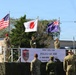 This image depicts a military ceremony for 'Orient Shield 2026,' a joint training exercise involving the United States, Japan, and Australia. Three soldiers are raising their respective national flags on a raised platform, with a banner displaying the exercise name in both English and Japanese. The event appears to take place at a military training area in Hokkaido, Japan, as indicated by the banner text.