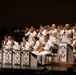 A United States Navy band performs on a concert hall stage, with musicians dressed in traditional white Navy dress uniforms. The ensemble includes both brass and woodwind sections, with trombones and trumpets visible in the back row and saxophones in the front. Music stands bearing the United States Navy Band insignia — featuring an eagle emblem — are positioned in front of the seated musicians.