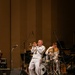 A U.S. Navy musician in dress white uniform performs a trumpet solo on a concert hall stage. Behind him, a second Navy musician in matching dress whites plays a drum kit. An audience is visible in the foreground, seated in theater-style chairs.