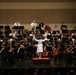 A large orchestra is performing on a concert hall stage, conducted by a person in a white outfit standing on a podium with their back to the camera. The ensemble appears to be a combined orchestra featuring both civilian musicians dressed in black and military personnel in white uniforms. The performance takes place in a modern, acoustically designed concert hall with a full audience visible in the foreground.