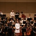 A naval officer in white dress uniform stands on a conductor's podium on a concert hall stage, facing an audience. She is surrounded by a large orchestra comprising both military personnel in white uniforms and civilian musicians in black attire. The setting appears to be a formal joint military-civilian orchestral concert performance.