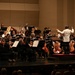 A full symphony orchestra is performing on a concert hall stage, led by a conductor in a white jacket who is facing away from the camera with arms raised. The musicians, dressed predominantly in black formal attire, are arranged in the traditional orchestral semicircle with string sections in the foreground and brass/woodwind sections in the background. The venue appears to be a modern concert hall with warm beige acoustic wall panels.