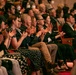 A diverse audience is seated in a theater or concert hall with red carpeting and wooden chairs, enthusiastically applauding a performance. The crowd spans multiple rows and appears to include people of various ethnicities and ages. The atmosphere suggests a formal cultural or performing arts event.