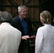 A group of five people are engaged in conversation in what appears to be a backstage or industrial-style venue. A tall man with gray hair wearing a navy blazer and blue shirt is the central figure, gesturing with his hands while speaking. The group includes people in both formal and casual attire, suggesting a professional or semi-formal gathering.
