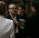 A group of people are engaged in conversation in what appears to be an indoor venue with wooden shelving or storage in the background. The central figure is an older East Asian man wearing glasses and a dark blazer, speaking with others around him. The setting suggests a formal or semi-formal gathering, possibly a reception or post-event meet-and-greet.