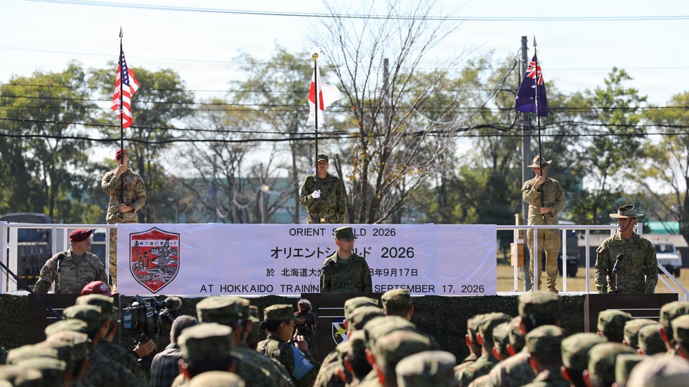 A multinational military ceremony titled 'Orient Shield 2026' is taking place at Hokkaido Training Area on September 17, 2026. Three flag bearers stand on an elevated stage holding the flags of the United States, Japan, and Australia, while military officers address assembled troops. The event appears to be an opening or closing ceremony for a joint military exercise involving at least three nations.