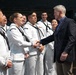 A civilian man in a dark blazer and blue shirt is shaking hands with a female U.S. Navy sailor in dress white uniform. Several other sailors in dress white uniforms are lined up in the background, suggesting a formal meet-and-greet or recognition event. The setting appears to be an indoor facility, possibly a hangar or gymnasium.