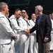 A civilian man in a dark blazer and blue shirt is shaking hands with a U.S. Navy sailor dressed in white dress uniform. The group of sailors, all wearing standard white Navy dress uniforms with black neckerchiefs, are lined up and appear to be in good spirits. The setting appears to be an indoor venue or ship deck during what looks like an official meet-and-greet or recognition event.