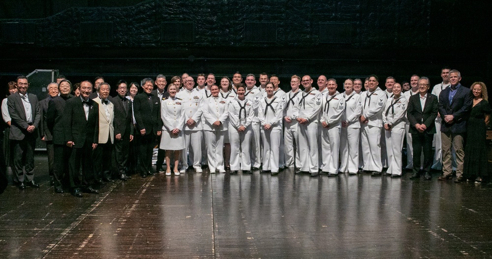 A large group photograph taken on what appears to be a stage or performance venue, featuring a mix of U.S. Navy personnel in white dress uniforms and civilians in formal attire. The group includes both men and women, with a notable presence of individuals of Asian descent among the civilian contingent on the left side. This appears to be a formal commemorative or diplomatic event involving military and civilian dignitaries.