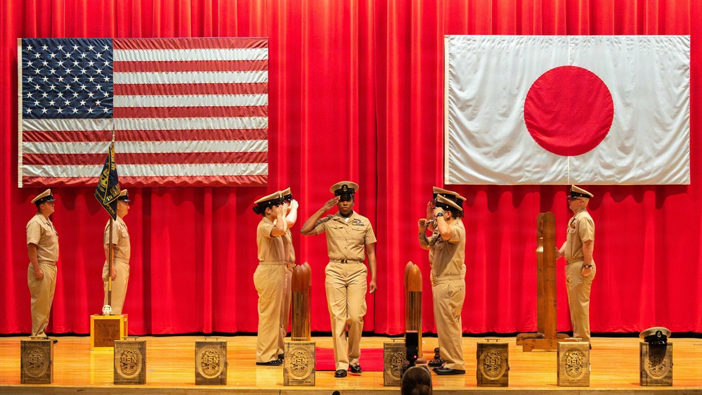A formal U.S. Navy military ceremony is taking place on a stage, with personnel in tan/khaki service uniforms rendering salutes. The backdrop features large American and Japanese flags against red curtains, indicating a bilateral or joint military event likely held in Japan. Several wooden podiums or stands bearing U.S. Navy (USN) insignia are arranged across the stage.