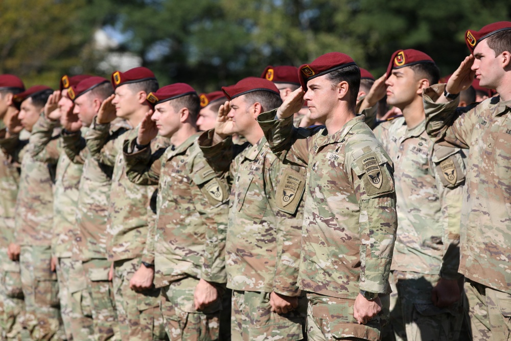 A large formation of U.S. Army soldiers wearing maroon berets and MultiCam (OCP) camouflage uniforms are rendering a salute in unison during a formal military ceremony. The soldiers display various patches including Airborne and Ranger tabs visible on the foreground soldier's left shoulder. The setting appears to be an outdoor parade field with trees visible in the background.