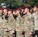 A large formation of U.S. Army soldiers wearing maroon berets and MultiCam (OCP) camouflage uniforms are rendering a salute in unison during a formal military ceremony. The soldiers display various patches including Airborne and Ranger tabs visible on the foreground soldier's left shoulder. The setting appears to be an outdoor parade field with trees visible in the background.